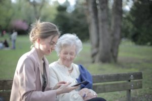 A younger woman using a smartphone with an elderly woman on a park bench.